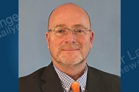 Bald man with glasses and a short beard, wearing a plaid shirt, orange tie, and jacket, smiling against a light blue background.