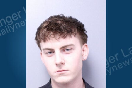 Mugshot of a young man with short brown hair, blue eyes, and silver stud earrings against a light background.