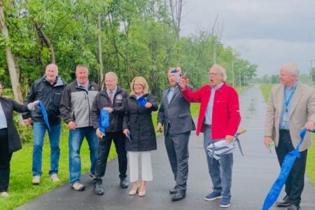 A group of local officials cut a blue ribbon to dedicate a new paved trail surrounded by trees.