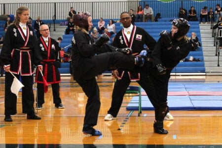 Two martial artists in black uniforms and protective gear spar on a gym floor, with one kicking and officials observing a competition.