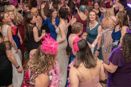 Women in various dresses and festive accessories dance and mingle at a vibrant indoor celebration.