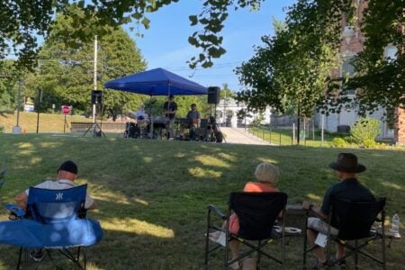 A band performs an outdoor concert under a blue canopy on a sunny lawn, with attendees watching near a large brick building.
