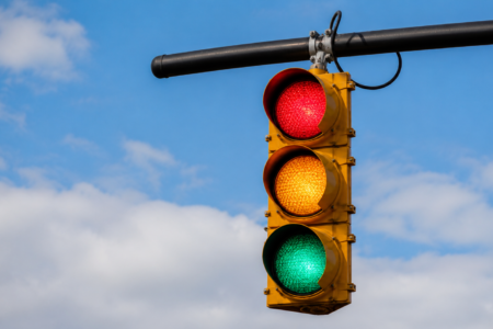 Yellow traffic light with red, yellow, and green signals all lit at once, against a blue sky with white clouds.