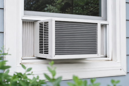 A white window air conditioner is installed in a window of a light blue house, with green leaves in the foreground.