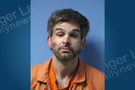 Man with greying hair and beard wearing an orange jumpsuit, looking forward against a blue background.