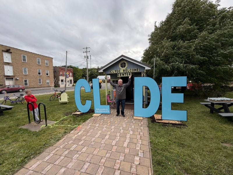 Man with raised arms stands between giant 'CLYDE' letters at the Clyde Welcome Center entrance, a boy nearby.
