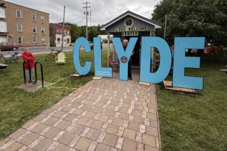 Man and boy pose by large blue "CLYDE" letters and the Clyde Welcome Center on a grassy area.
