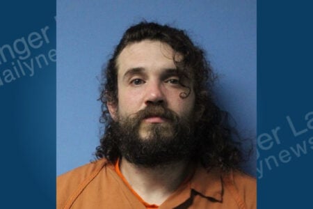 A man with a full beard, long curly hair, and an orange uniform looks directly at the camera against a blue background.