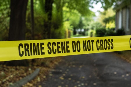 Yellow "CRIME SCENE DO NOT CROSS" tape stretched across a residential street with trees and a house in the background.