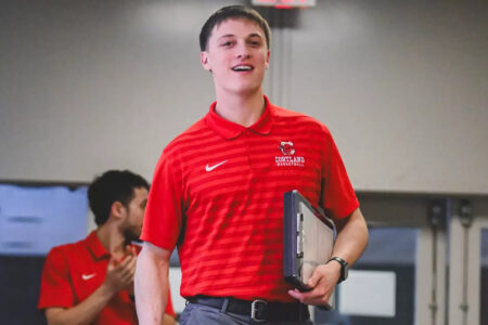 Cortland Basketball staff member in a red polo shirt smiles while walking indoors, holding a clipboard.