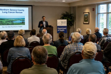A speaker presents a "Northern Seneca County Lung Cancer Review" to an audience, with a NYS Department of Health banner nearby.