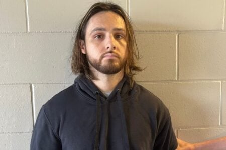 Bearded man with medium-length brown hair in a black hoodie stands against a beige cinder block wall.