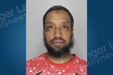 Man with a beard, short hair, and facial tattoos, wearing a red splatter shirt, looks forward in a mugshot.