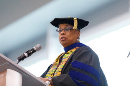 A Black woman in a graduation gown and kente stole speaks into a microphone at a podium.