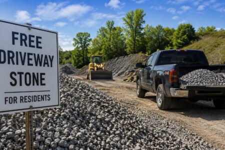 A "Free Driveway Stone For Residents" sign at a gravel distribution site, with a pickup truck loaded with stone and a loader.
