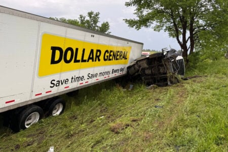 Dollar General semi-truck with overturned cab and leaning trailer in tall grass off a road.