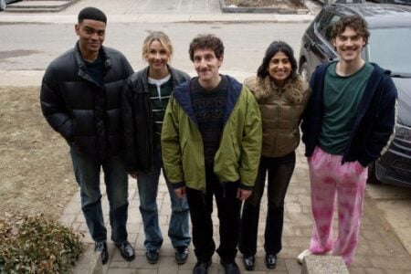 Five smiling young adults pose on a residential driveway, wearing winter jackets.