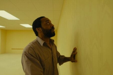 Bearded man in brown shirt looks up with open mouth, hand pressed on a pale yellow wall in a room with fluorescent lights.