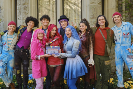 Nine smiling cast members from Descendants: The Rise of Red pose with a clapboard outside a stone building.