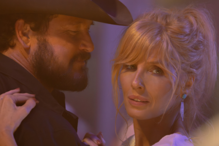 A blonde woman with an intense gaze and a bearded man in a cowboy hat embrace under warm, purple lighting.