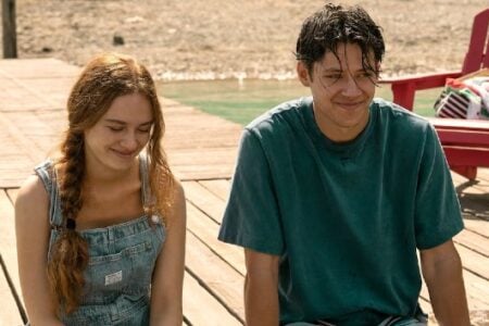 Two young people, a woman in overalls and a man in a t-shirt, relax and smile on a wooden dock by a Finger Lakes shore.