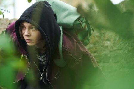Young woman in black hoodie and green backpack crouches, peering cautiously through green foliage near a stone wall.