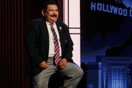 Smiling man in blazer and tie poses on stage with a Hollywood sign and Griffith Observatory backdrop.