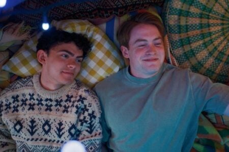 Two smiling young men lie in a cozy fort under blue string lights, one holding a camera for a selfie.