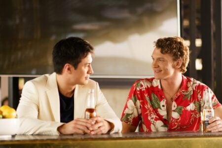 Two smiling men chat at a bar counter, one in a cream blazer, the other in a red Hawaiian shirt with a Corona.