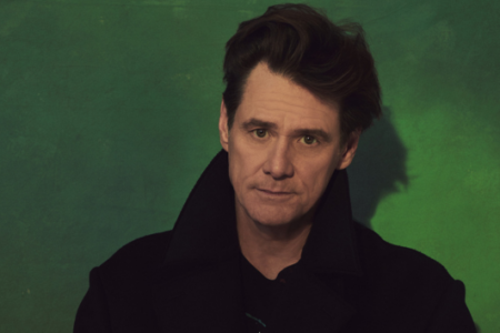 Jim Carrey, wearing a dark coat, looks directly at the camera with a serious expression against a green background.