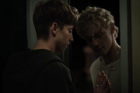 Two young people with brown and blonde hair press their hands and faces against a window screen in a dark scene.
