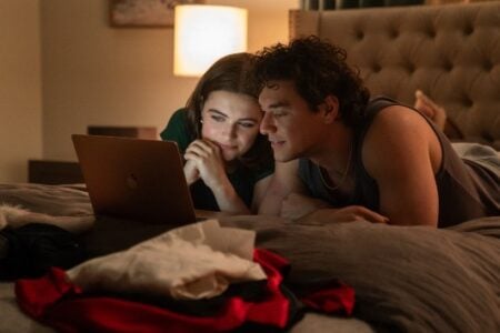 A young man and woman lie on their stomachs in bed, intently watching a laptop screen in a warmly lit bedroom.