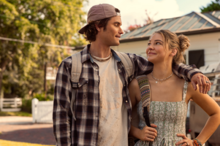 Smiling young man in a plaid shirt with arm around woman in a sundress, looking at each other in front of a house.