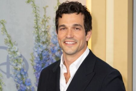 Man with dark curly hair and a white shirt under a dark jacket smiles against a light wall with blue floral patterns.