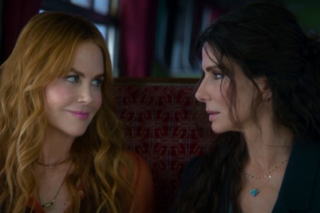 Nicole Kidman smiles at Sandra Bullock, who looks intently back, both seated in front of a red patterned wall.