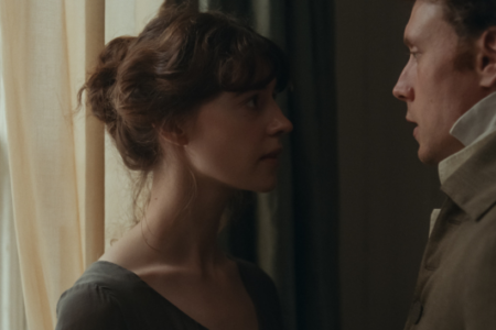 Serious young woman with brown hair gazes intently at a man in a beige coat, standing close indoors by a window.