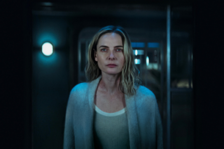 Blonde woman in a light cardigan with a mark on her neck looks directly forward in a dark, blue-lit hallway.