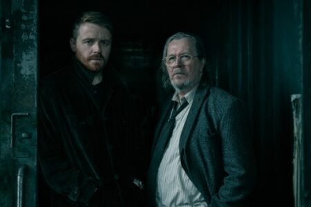 Two men, one younger with a beard and one older in glasses, stand in a dark shipping container doorway.