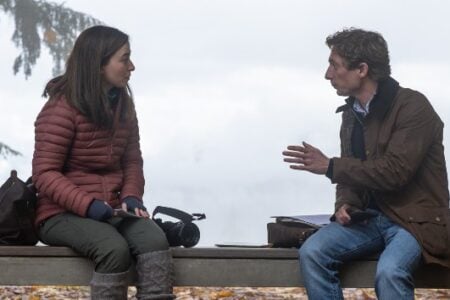 A woman in a red puffer jacket and a man in a brown jacket talk on an outdoor bench with trees and an overcast sky.