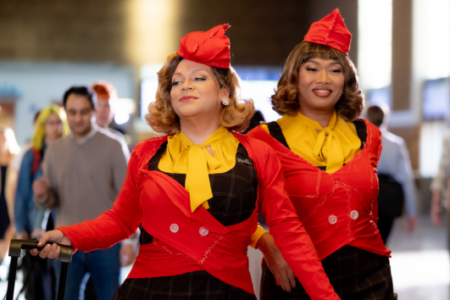 Two smiling drag queens in retro flight attendant costumes walk through a busy terminal; one holds a luggage handle.