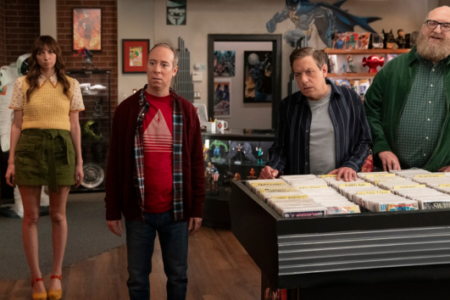 Four people, including a tall woman and a bearded man, stand attentively inside a comic book shop among display bins.