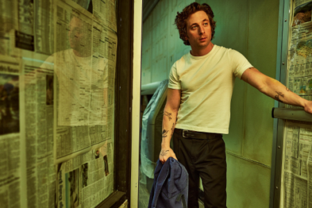 Jeremy Allen White in a white t-shirt leans on a rail in a green-lit room with walls covered in newspapers.