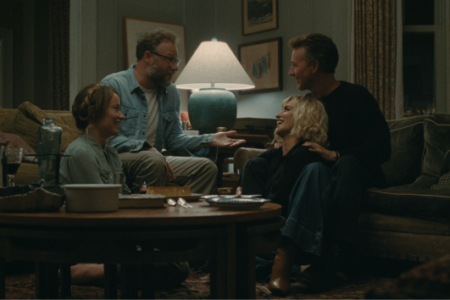 Four people laugh and converse in a living room, gathered around a coffee table with food and wine.