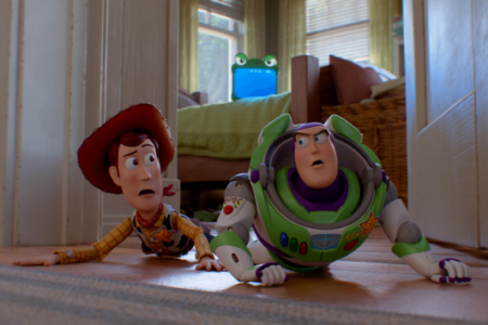 Woody and Buzz Lightyear crawl low on a wooden floor with startled expressions in a room.