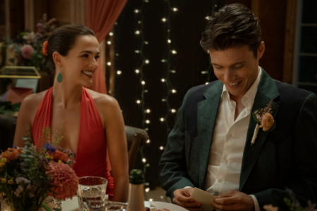 A smiling woman in red watches a man in a suit with a boutonnière read a card at a decorated table.