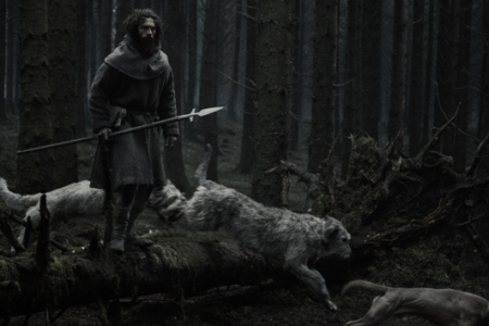 Bearded man in a hooded cloak stands on a mossy log, holding a spear, as two dogs run and leap in a dark forest.