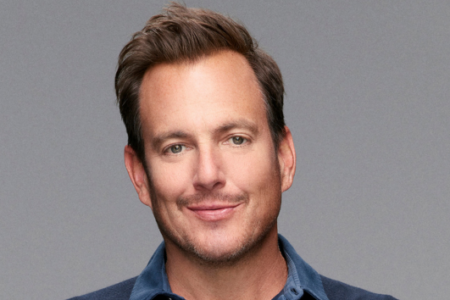 Will Arnett smiles with a light mustache and stubble, wearing a blue collared shirt against a grey background.