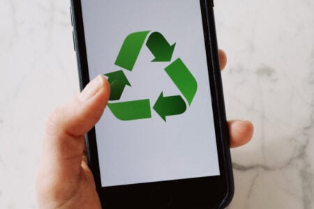 Hand holding a smartphone displaying a green recycling symbol on its screen over a white marble surface.