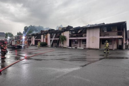 Firefighters respond to a severely burned two-story motel with a charred roof and smoke under a cloudy sky.