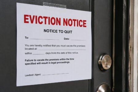 An eviction notice with "NOTICE TO QUIT" visible, taped to a dark door next to a brick wall.